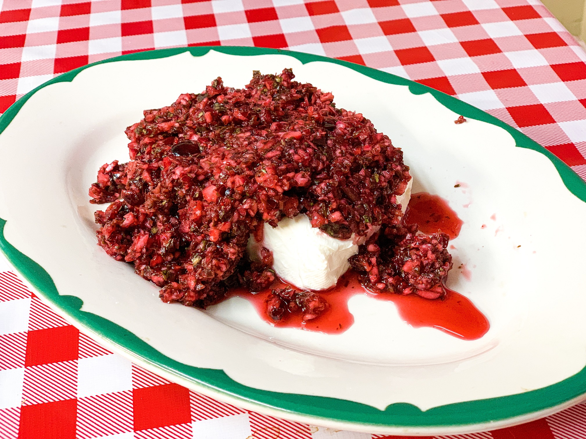 Cranberry salsa draped over a block of cream cheese. It is set on a white platter with a green rim on a red and white checkered tablecloth.